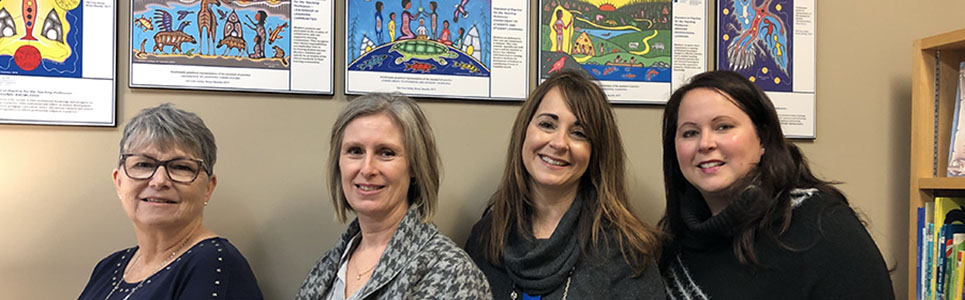 Four female adults standing along a wall with Indigenous artwork in the background